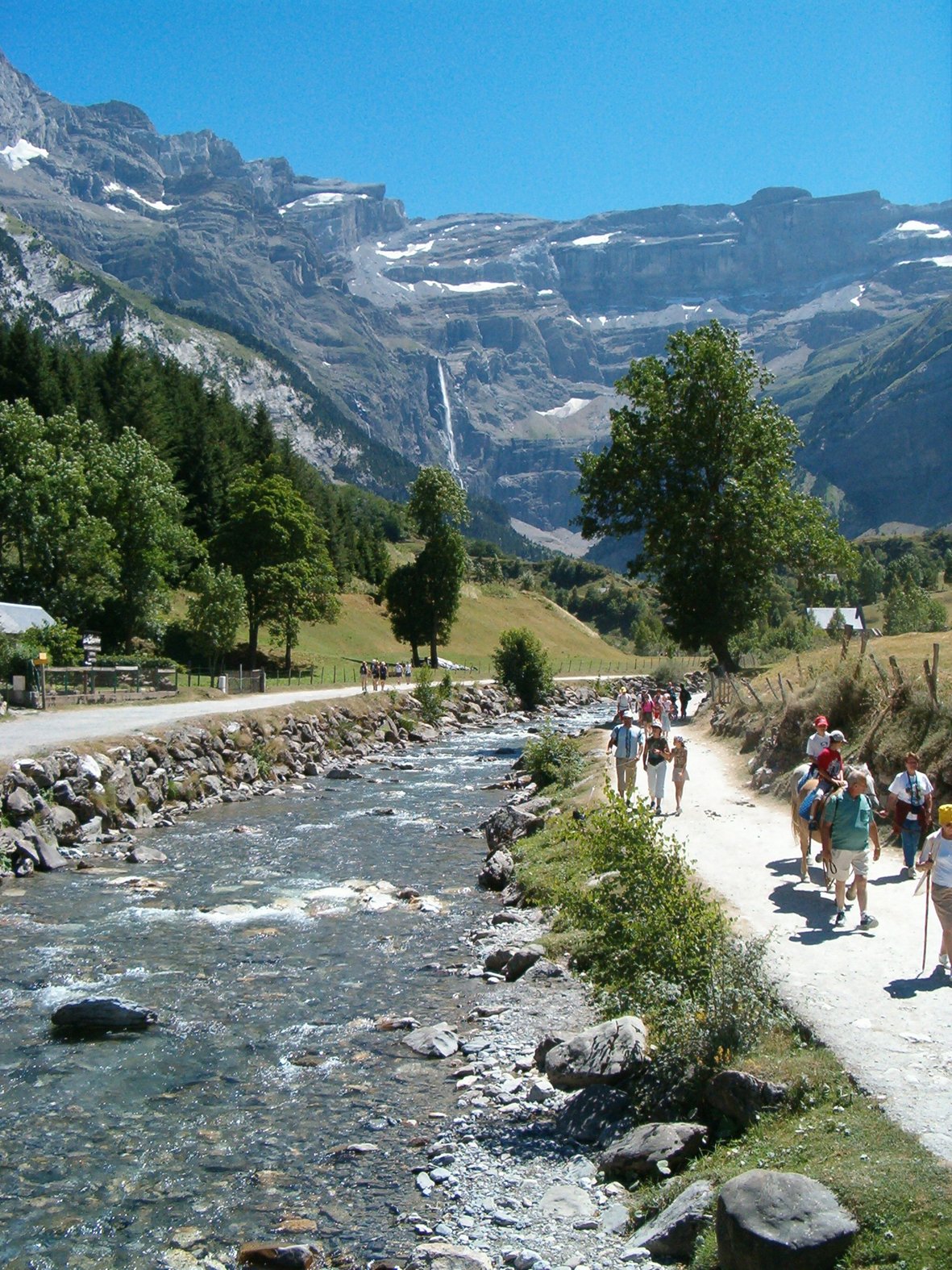 Randonneurs marchant le long d'une rivière dans une vallée alpine entourée d'arbres, de montagnes avec des glaciers, en plein jour.