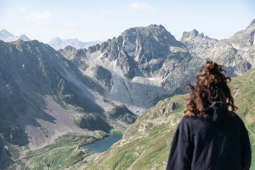 Une personne avec des cheveux bouclés et vêtue d'une veste noire regarde un paysage de montagnes escarpées et un lac en contrebas.