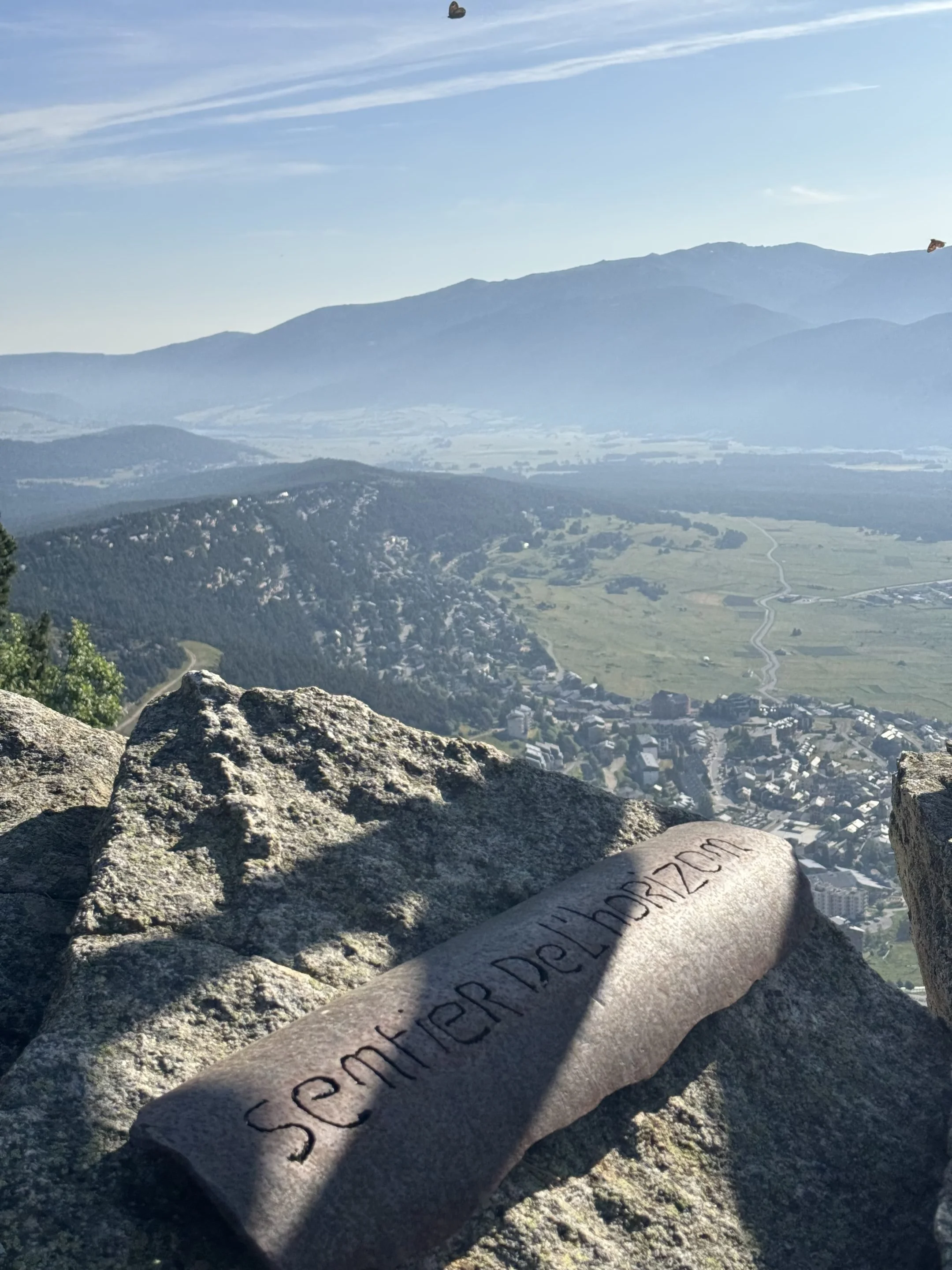 Vue montagneuse avec un sgotten de pierre gravé au premier plan, sur une plateforme en roche, surplombant une vallée avec des bâtiments et une route, ciel clair avec quelques nuages