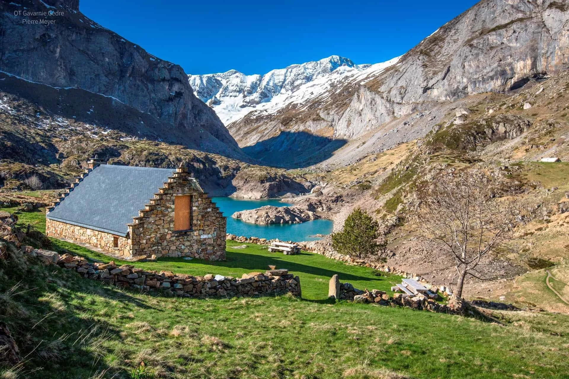 Une maison en pierre avec un toit en métal est située dans un paysage de montagne, avec un lac bleu à proximité, entourée de collines verdoyantes et de montagnes enneigées au fond, sous un ciel clair et bleu.