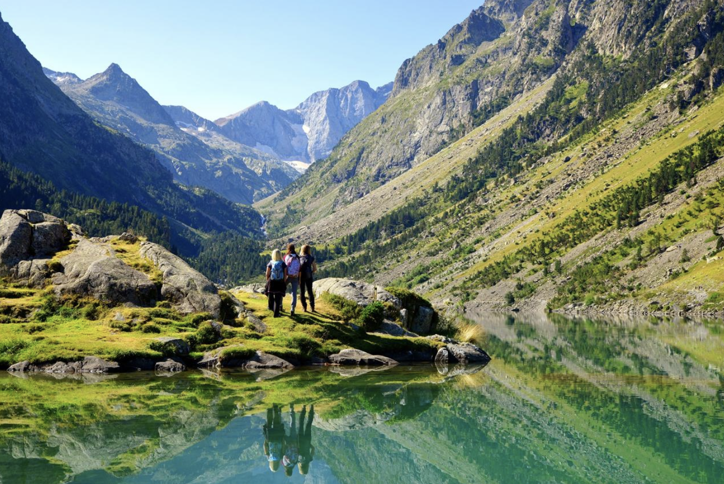 Trois randonneurs émergent d’un lac de montagne entouré de sommets enneigés, de forêts et de rochers.