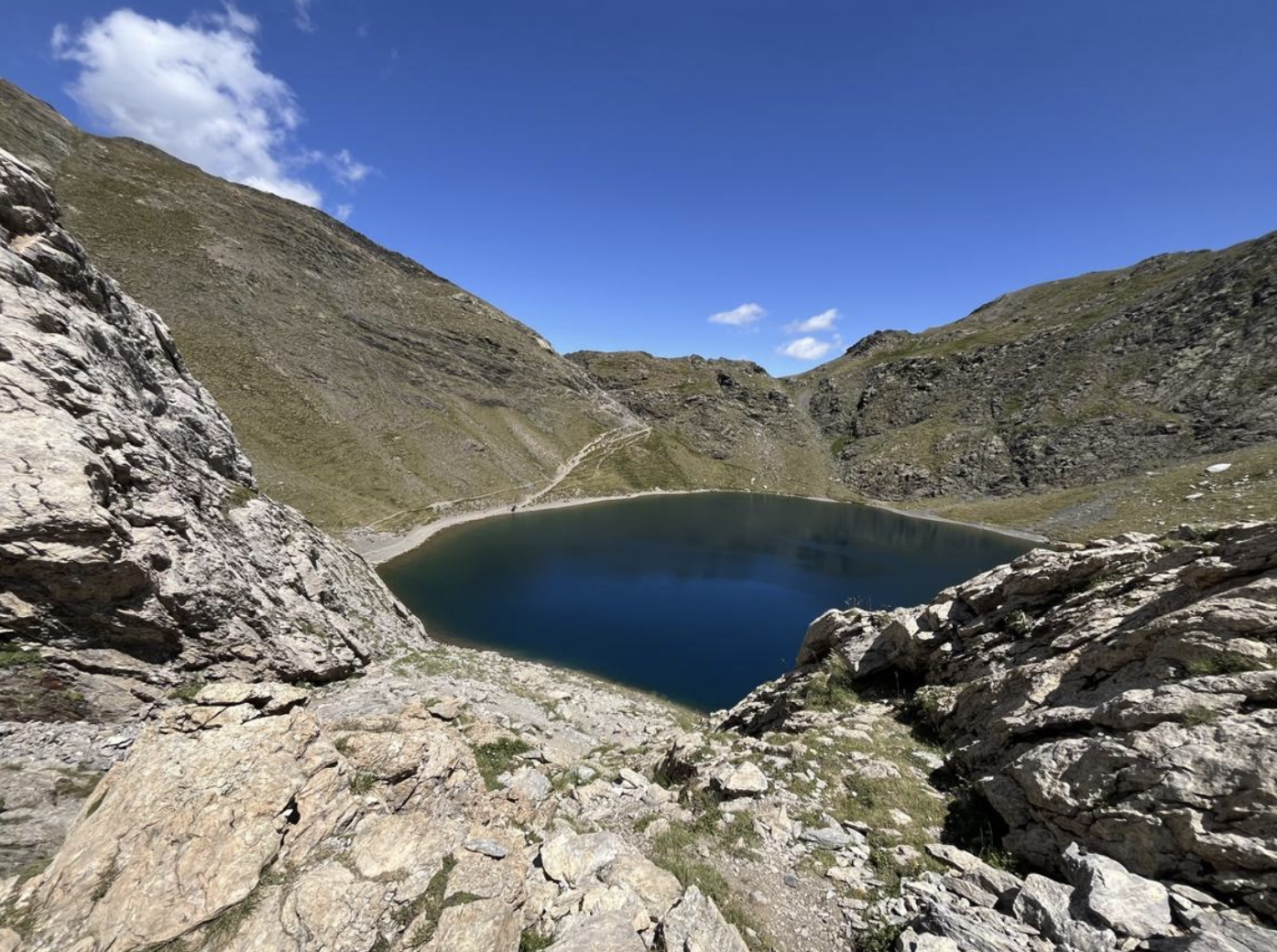 Lac de haute montagne entouré de rochers et de pentes rocheuses sous un ciel bleu clair avec quelques nuages