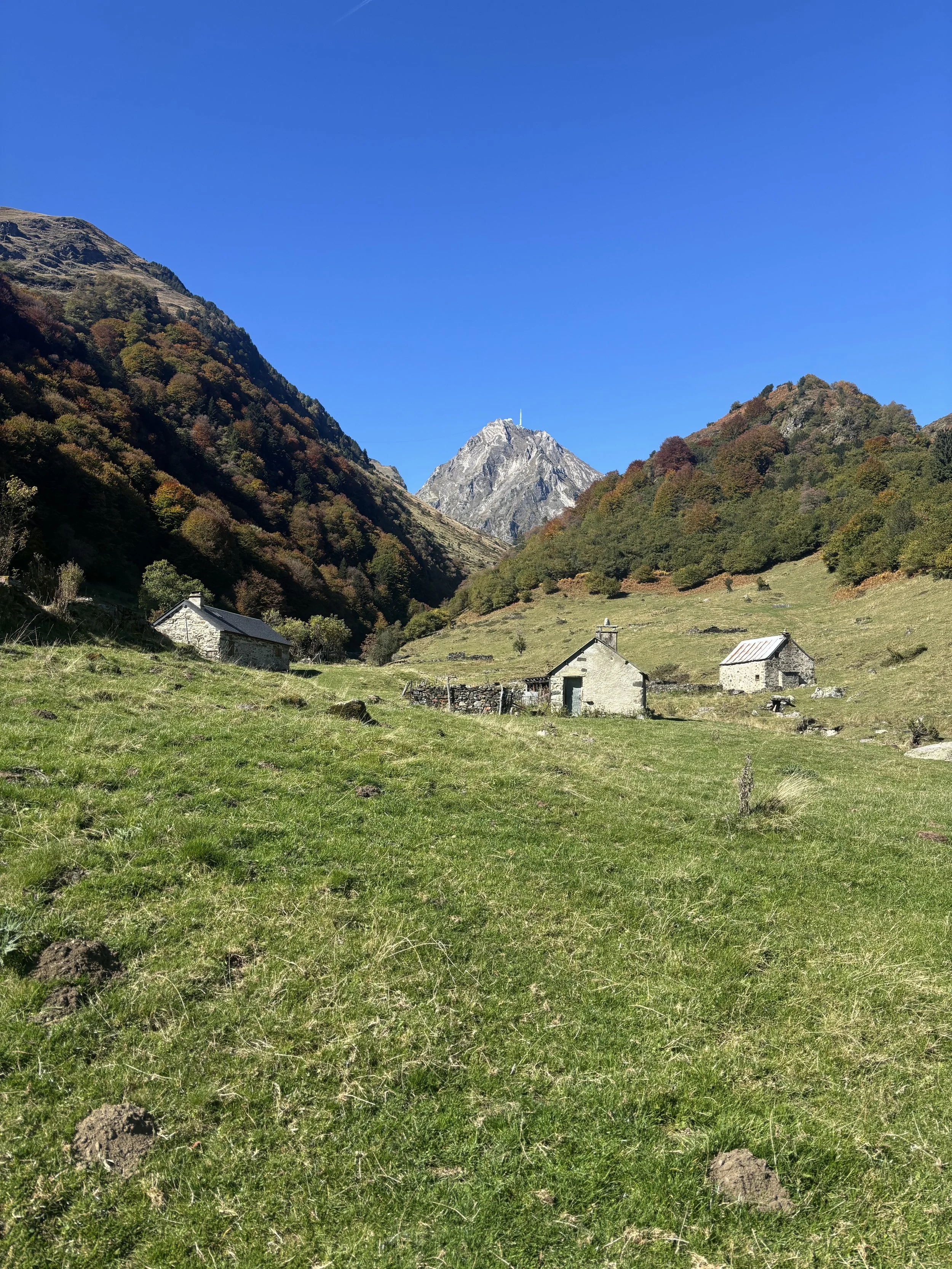 Paysage de montagne avec quelques maisons en pierre et un sommet enneigé dans le fond sous un ciel bleu clair.