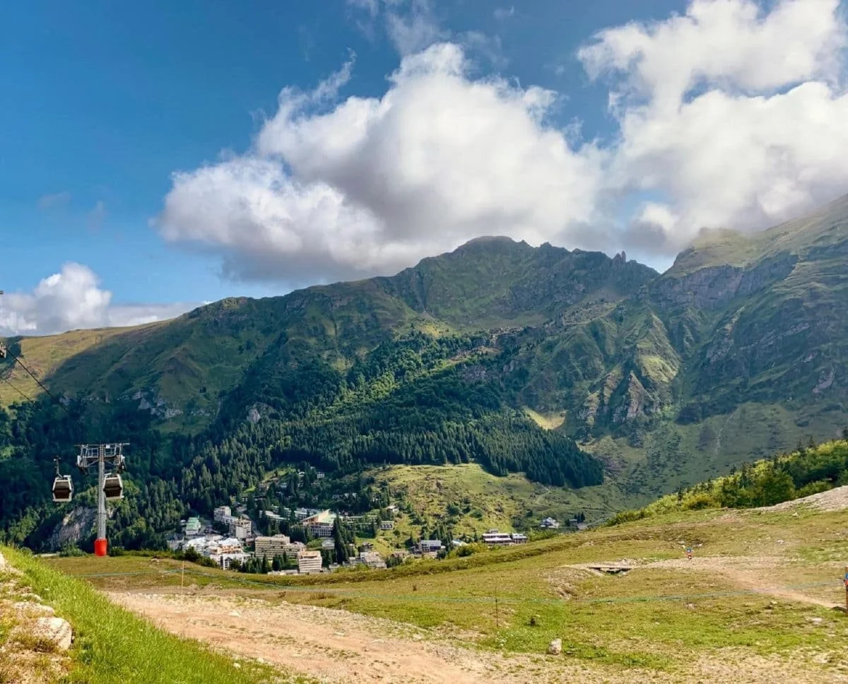 Paysage de montagnes verdoyantes sous un ciel partiellement nuageux, avec un village en contrebas et une télécabine sur la gauche.