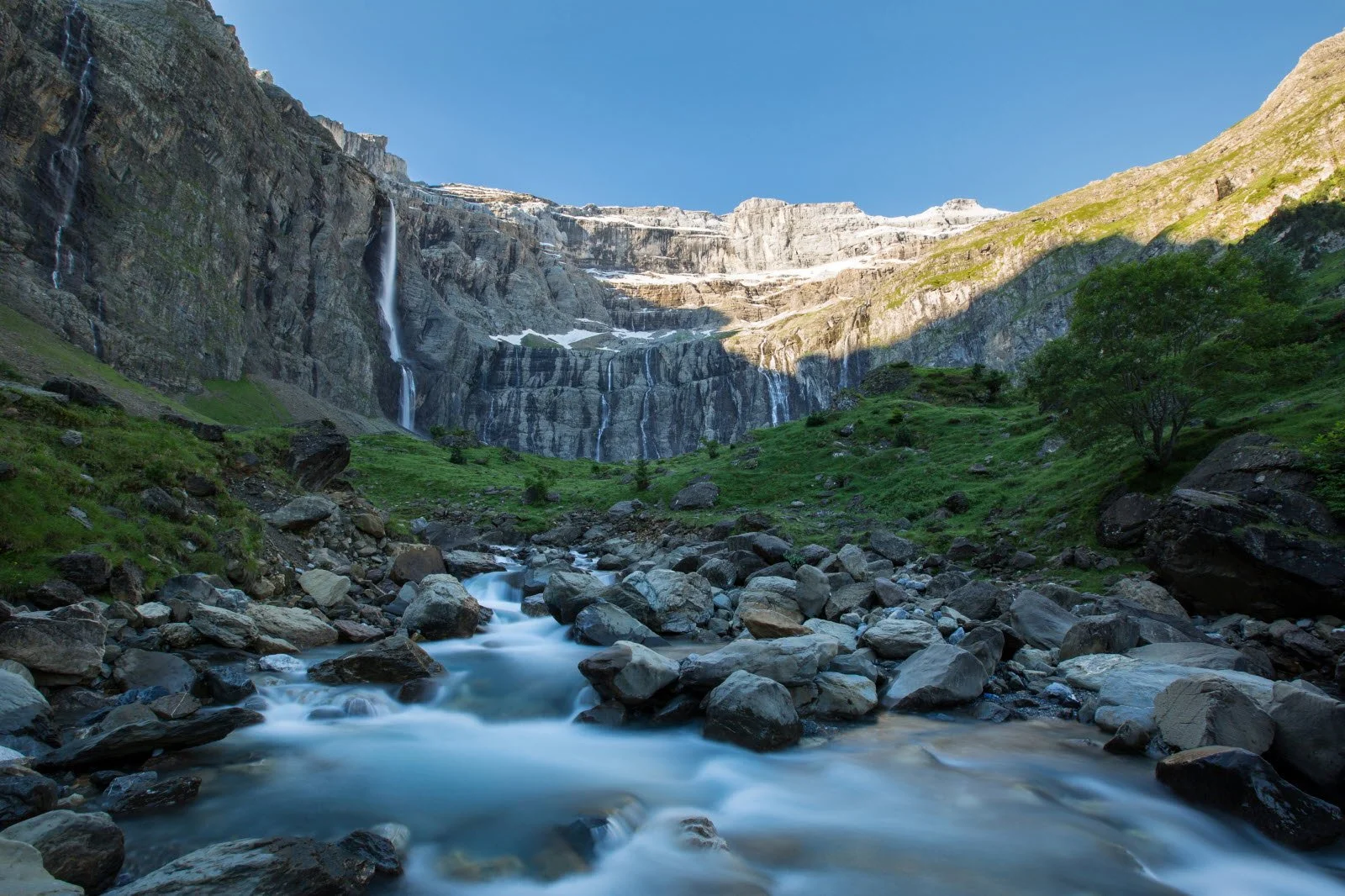 Vue d'une cascade dans une vallée herbeuse entourée de montagnes rocheuses sous un ciel bleu clair.