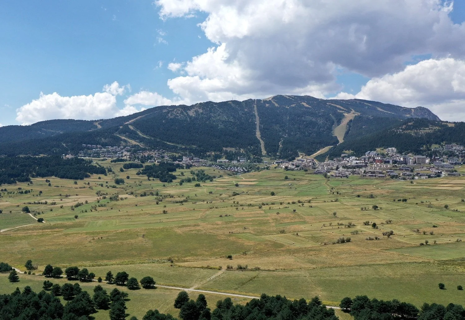 Paysage de montagnes avec un village, prairies et arbres sous un ciel partiellement nuageux.