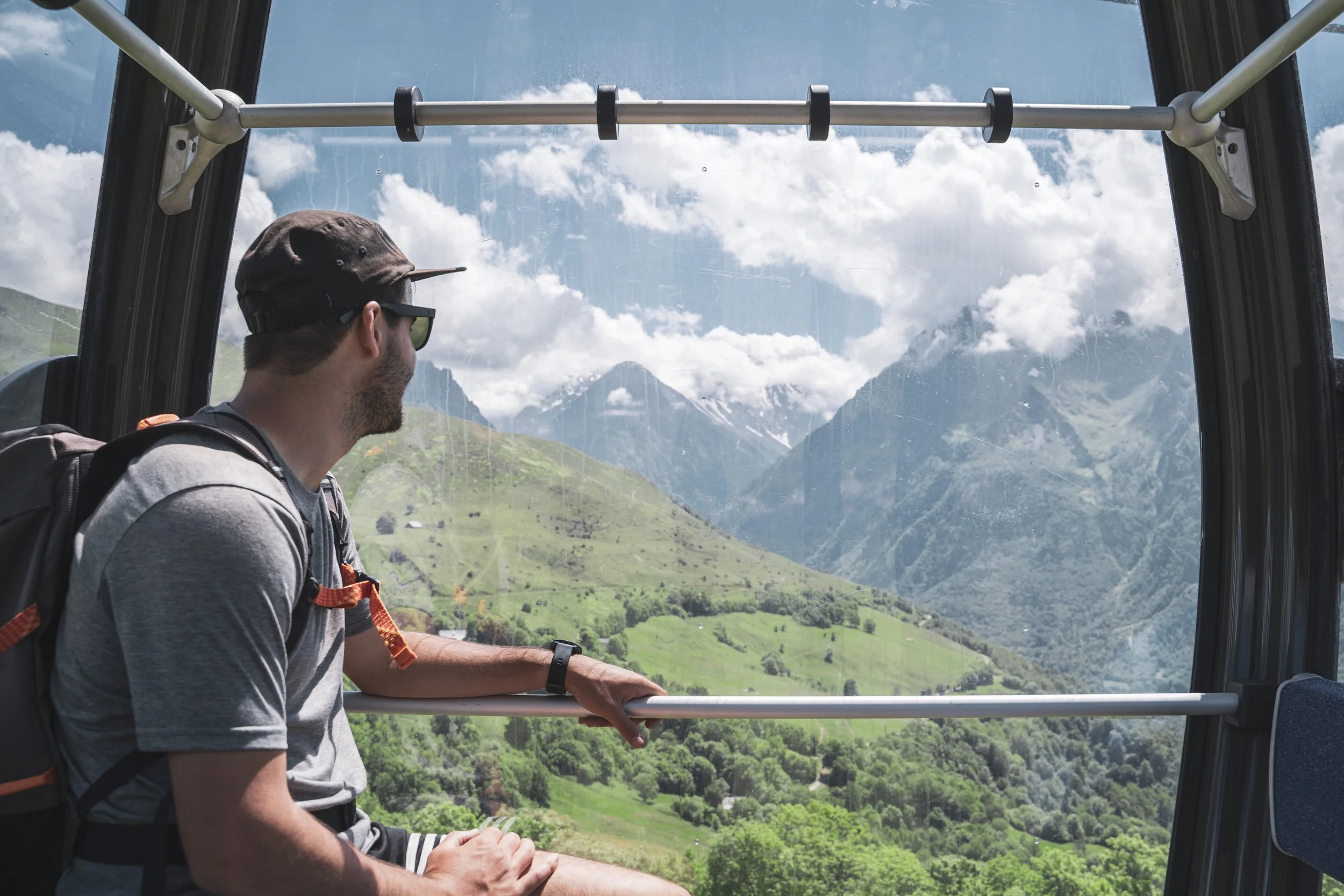 Un homme portant un chapeau et un sac à dos regarde par une fenêtre d'une cabine de téléphérique sur un paysage de montagnes, de vallées verdoyantes et un ciel bleu avec des nuages.
