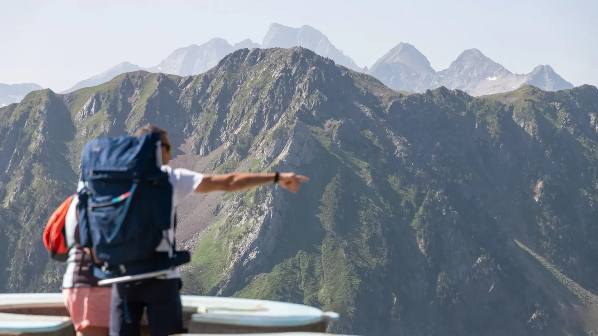 Un homme avec un sac à dos pointe vers une montagne dans un paysage de montagnes rocheuses avec peu de végétation.