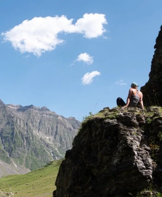 Une personne assise sur un rocher dans un paysage de montagne, avec un ciel bleu et quelques nuages blancs.