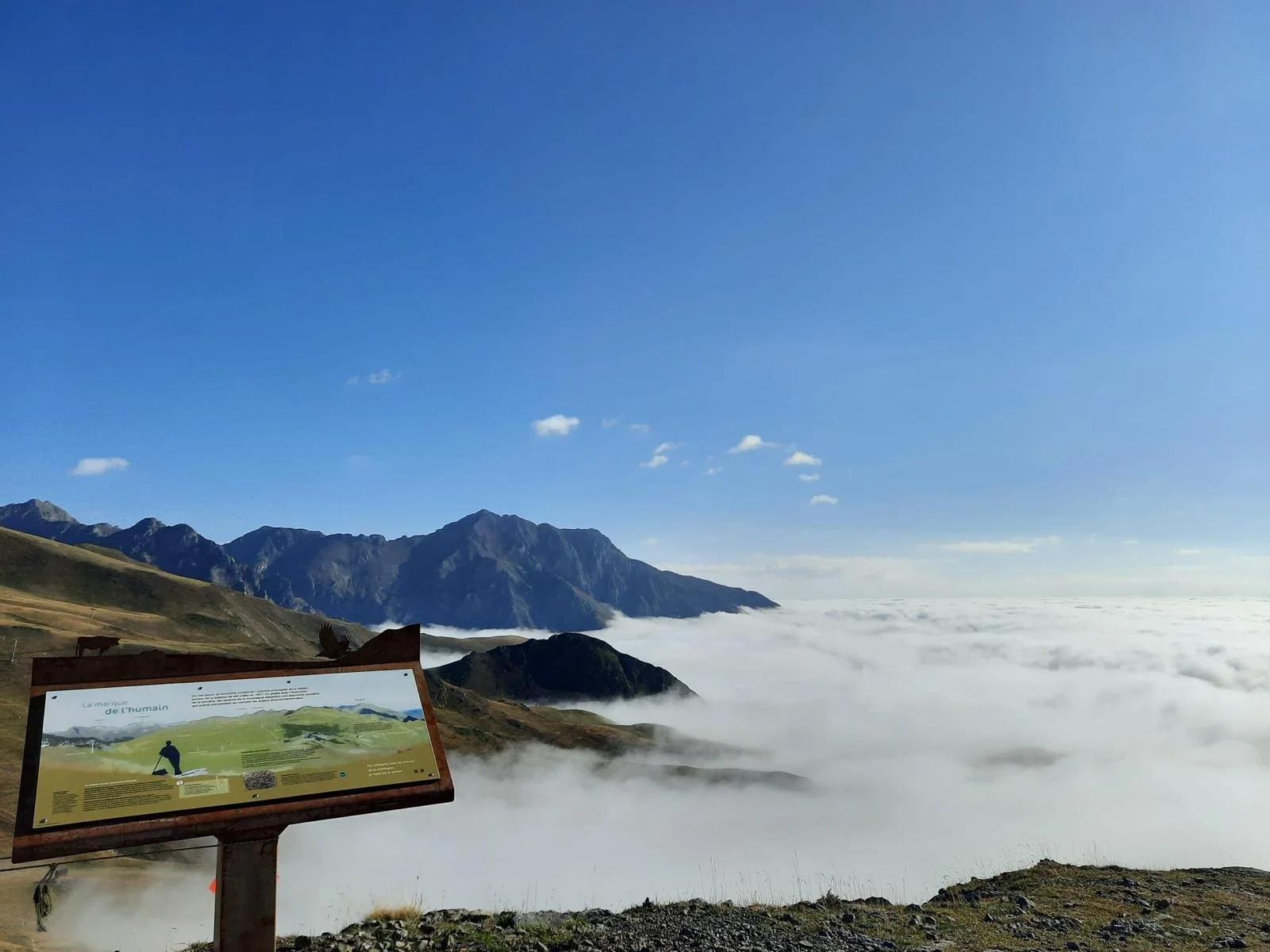 Paysage de montagnes avec ciel bleu, nuages, et brume ou nuages en bas, avec panneau d'information sur le site de l'Œil du massif.
