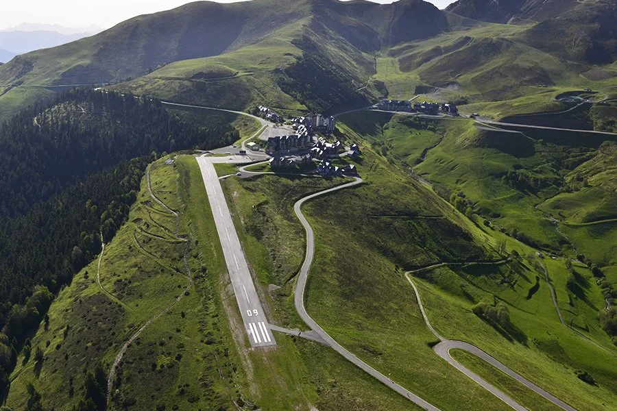 Vue aérienne d'une petite ville en altitude, entourée de montagnes verdoyantes, avec une zone de piste d'atterrissage commence par '50' et plusieurs routes sinueuses.