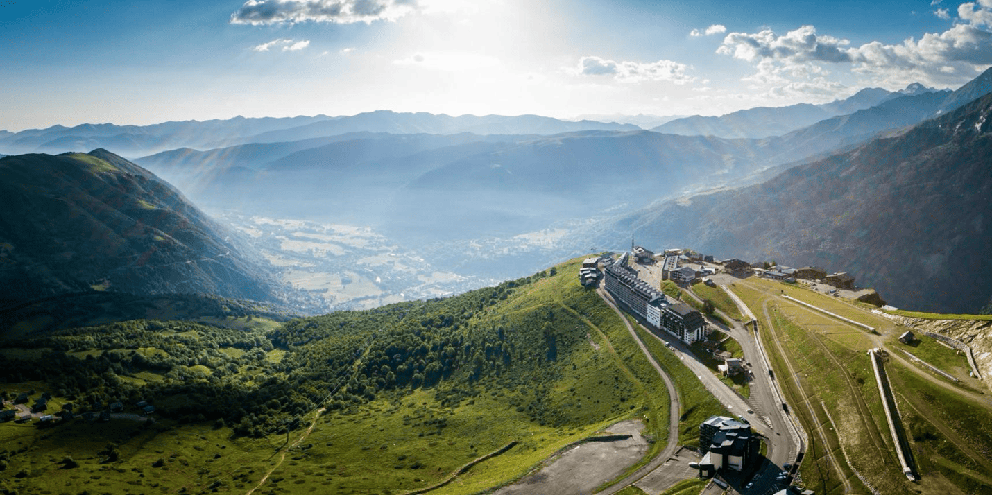 Paysage de montagnes avec un monastère ou un hôtel en haut d'une colline verte, avec un réseau de routes et des bâtiments environnants, sous un ciel partiellement nuageux.