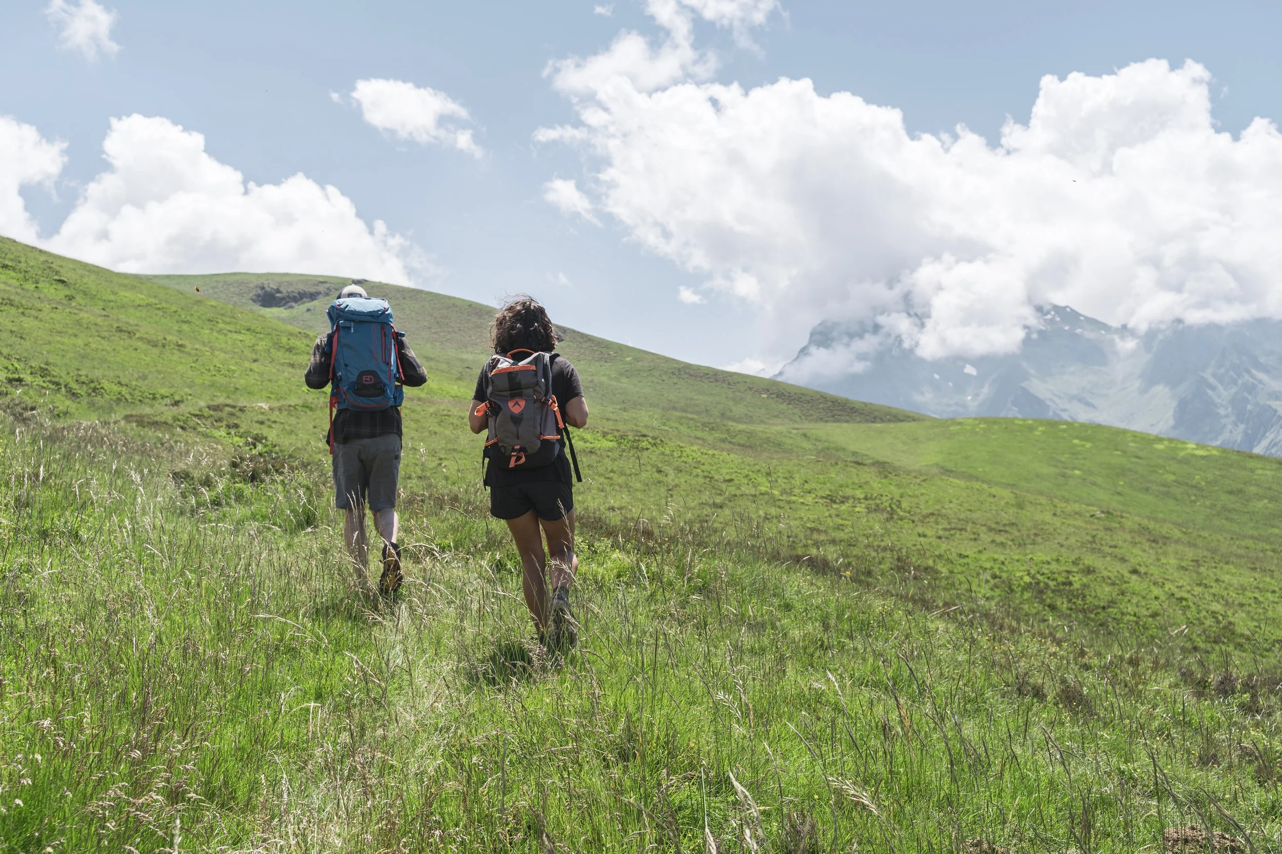 Deux personnes avec des sacs à dos faisant de la randonnée dans une vallée verdoyante entourée de montagnes sous un ciel partiellement nuageux.