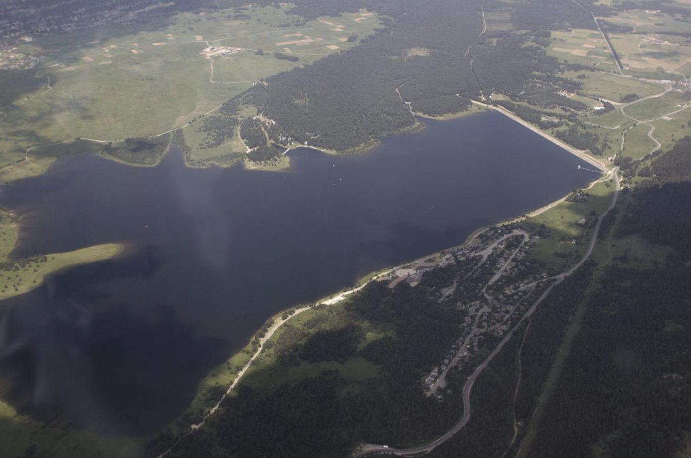 Vue aérienne d'un lac entouré de forêts, de champs agricoles et de petites zones résidentielles ou de villégiature, avec des routes qui le bordent.
