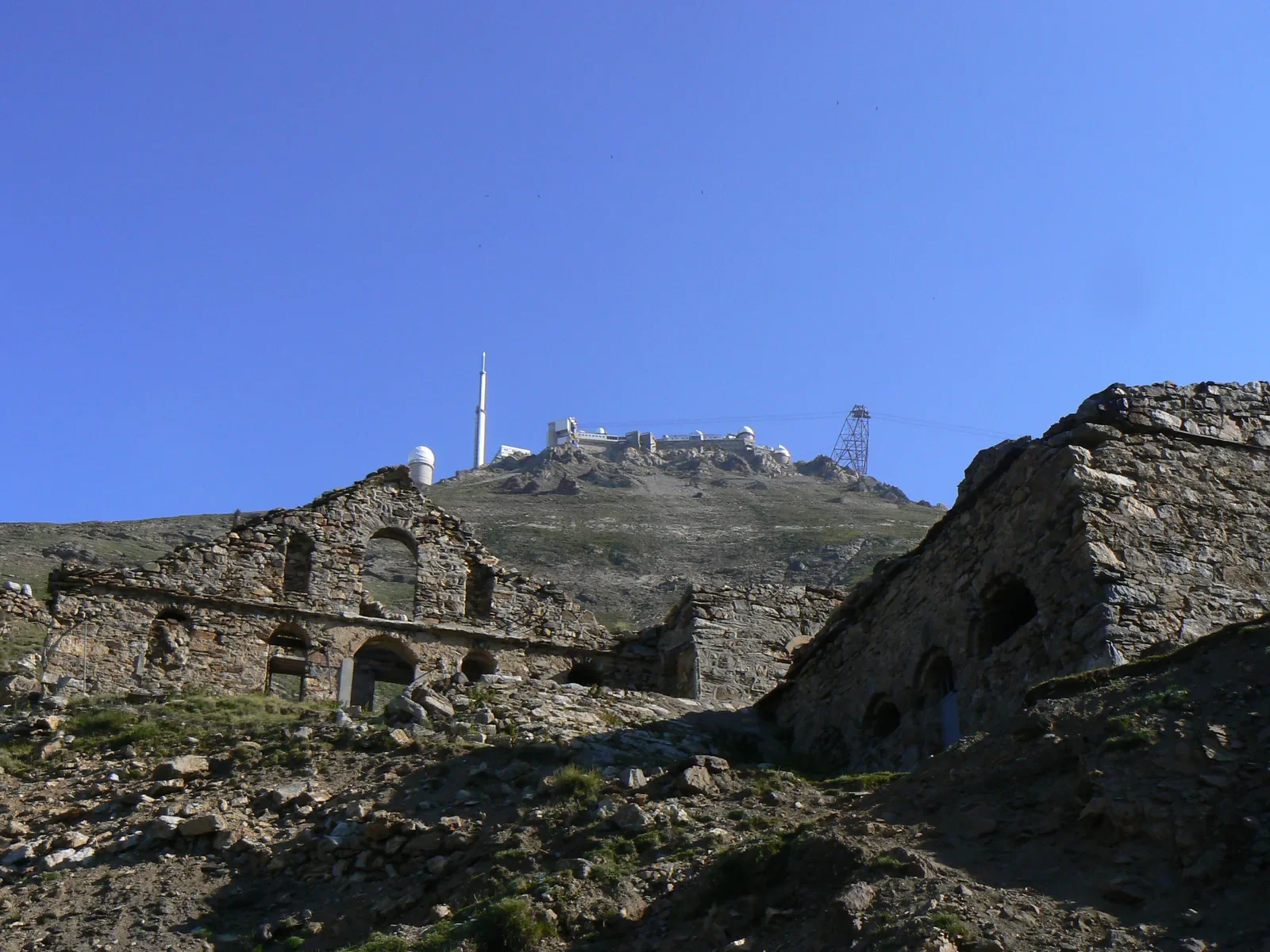 Ruines anciennes en pierre avec un fond de montagnes et d'installation de télécommunication ou d'observatoire en haut, sous un ciel bleu clair.