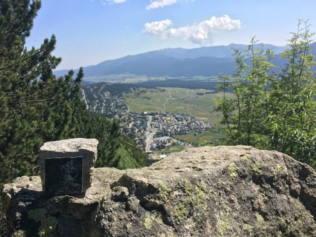 Paysage montagneux avec une ville en contrebas, arbres et rochers au premier plan, ciel bleu avec quelques nuages.