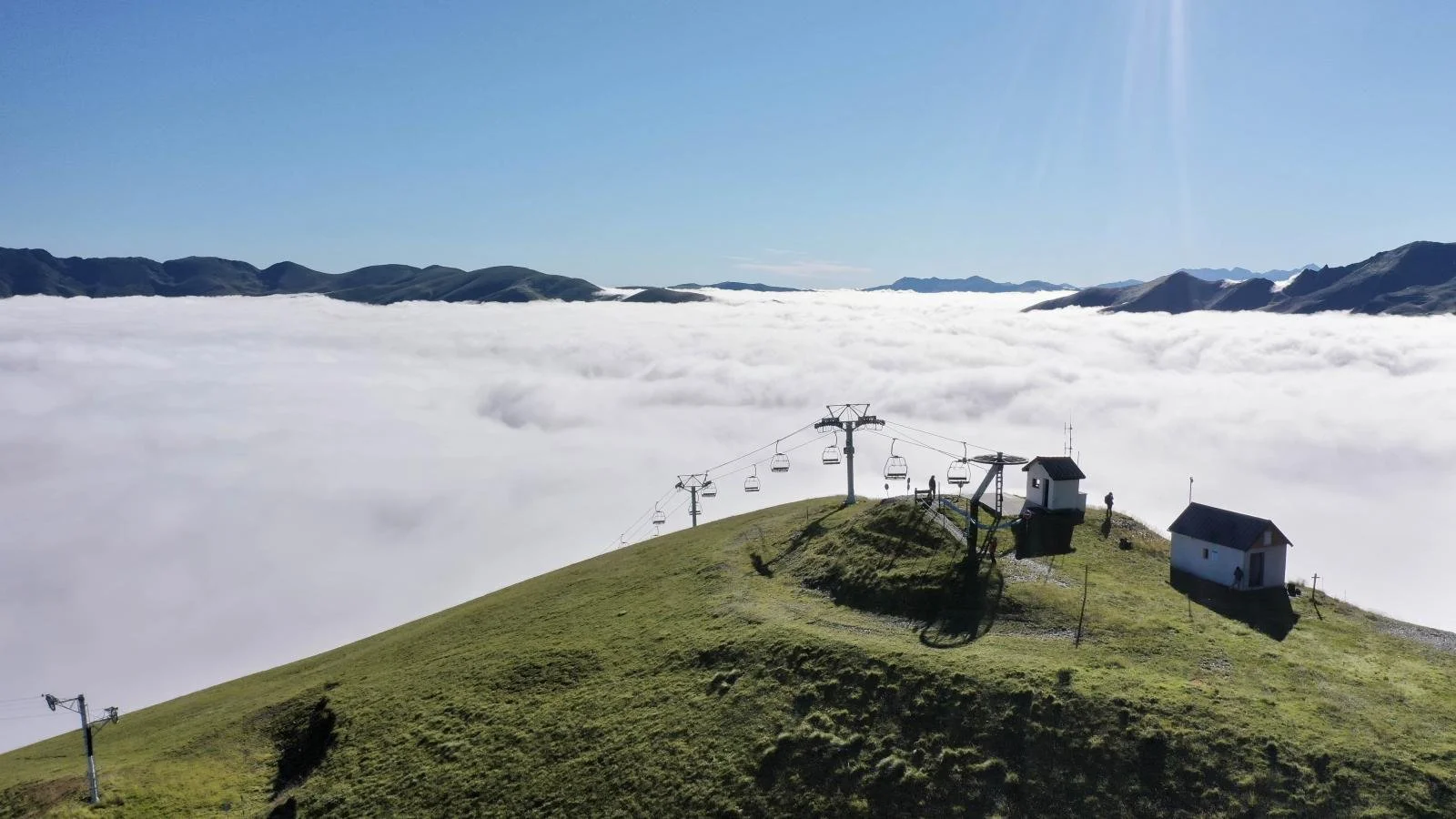 Montagne verdoyante avec un téléphérique et deux petites maisons, au-dessus d'une mer de nuages, sous un ciel clair et ensoleillé.