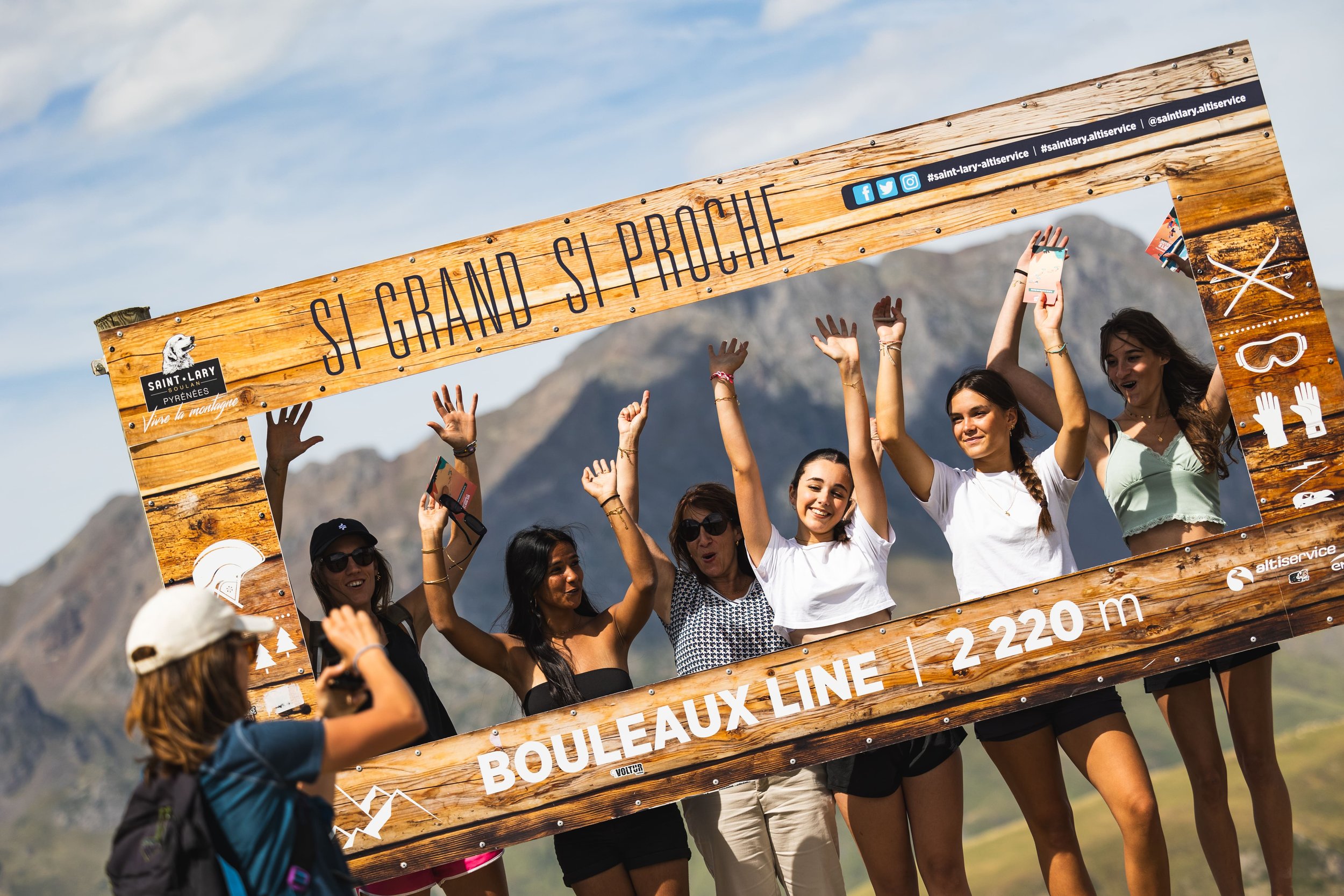 Groupe de jeunes femmes souriantes tenant une grande pancarte en bois avec l'inscription « SI GRAND SI PROCHE » dans un paysage montagneux en arrière-plan, lors d'une activité de plein air.