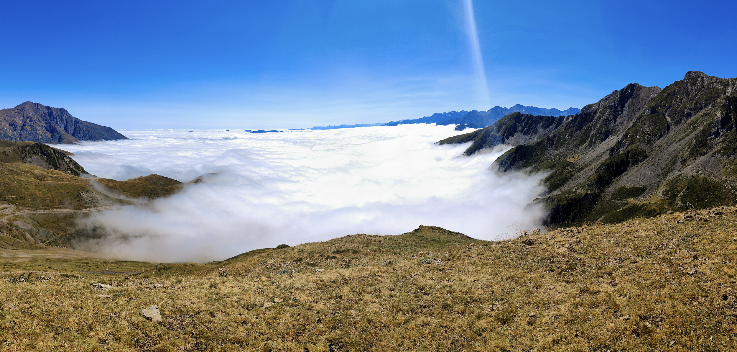 Vue panoramique de montagnes avec une mer de nuages au fond et un ciel bleu clair.