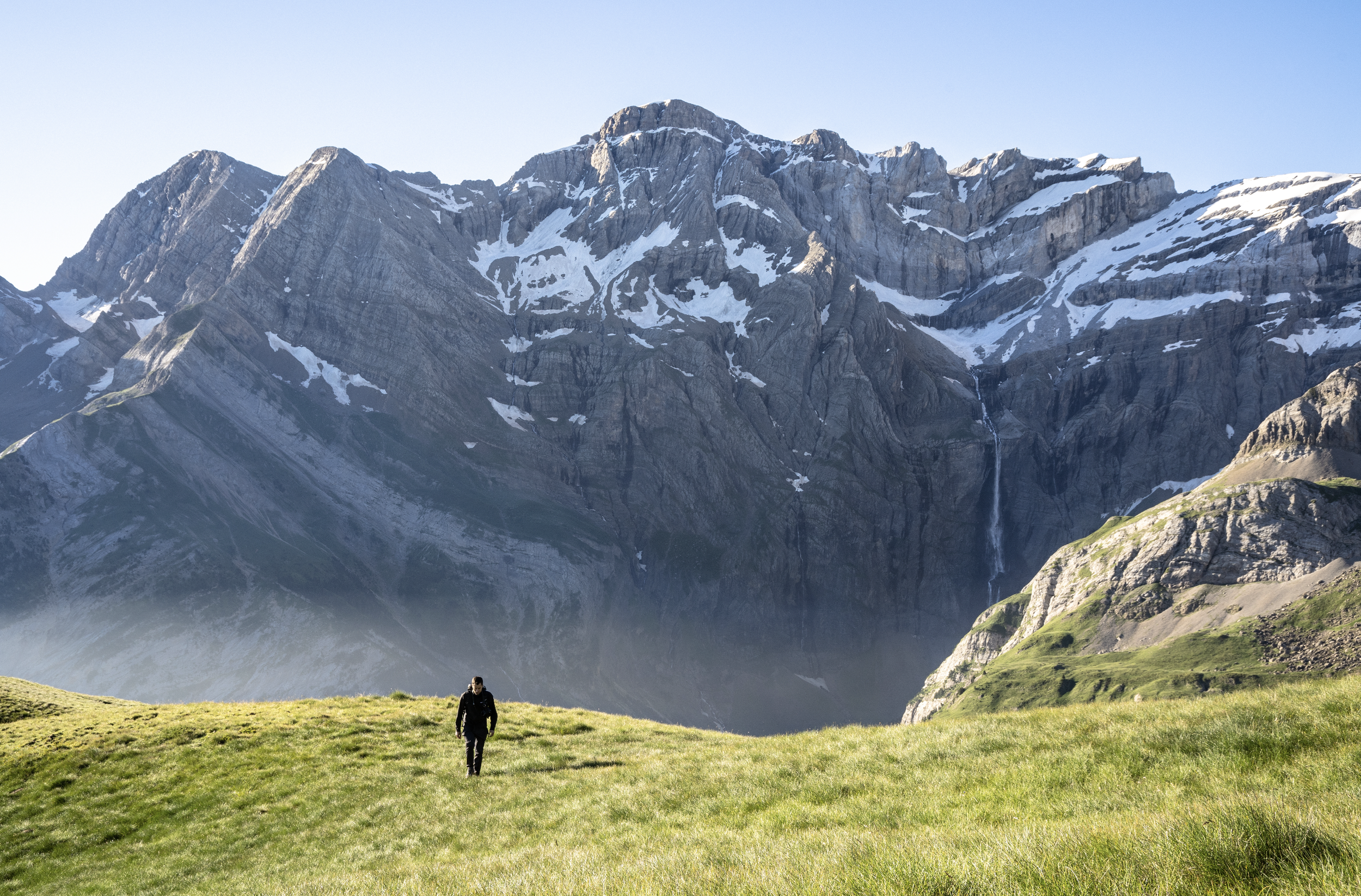Un homme marche dans une vallée verdoyante entourée de montagnes enneigées au loin, sous un ciel clair.