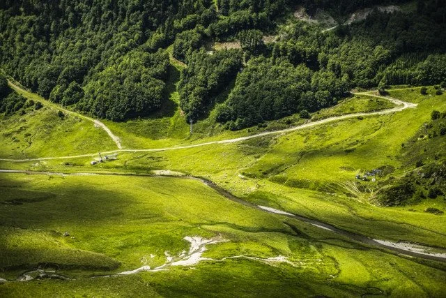 Paysage montagneux avec des pentes verdoyantes, des chemins serpentant à travers la vallée et des forêts denses en arrière-plan.