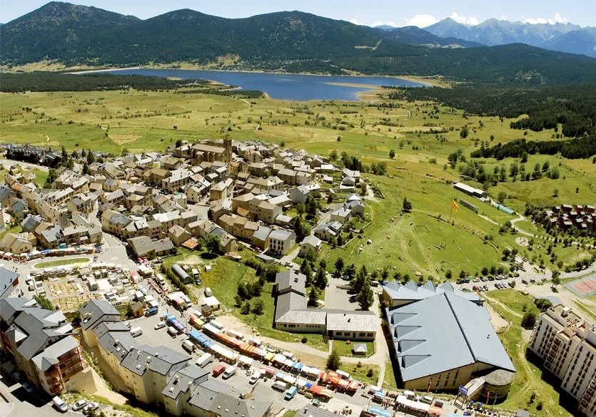 Vue aérienne d'un village entouré de montagnes, avec un lac au fond, un grand bâtiment, et une zone pavée avec des véhicules.