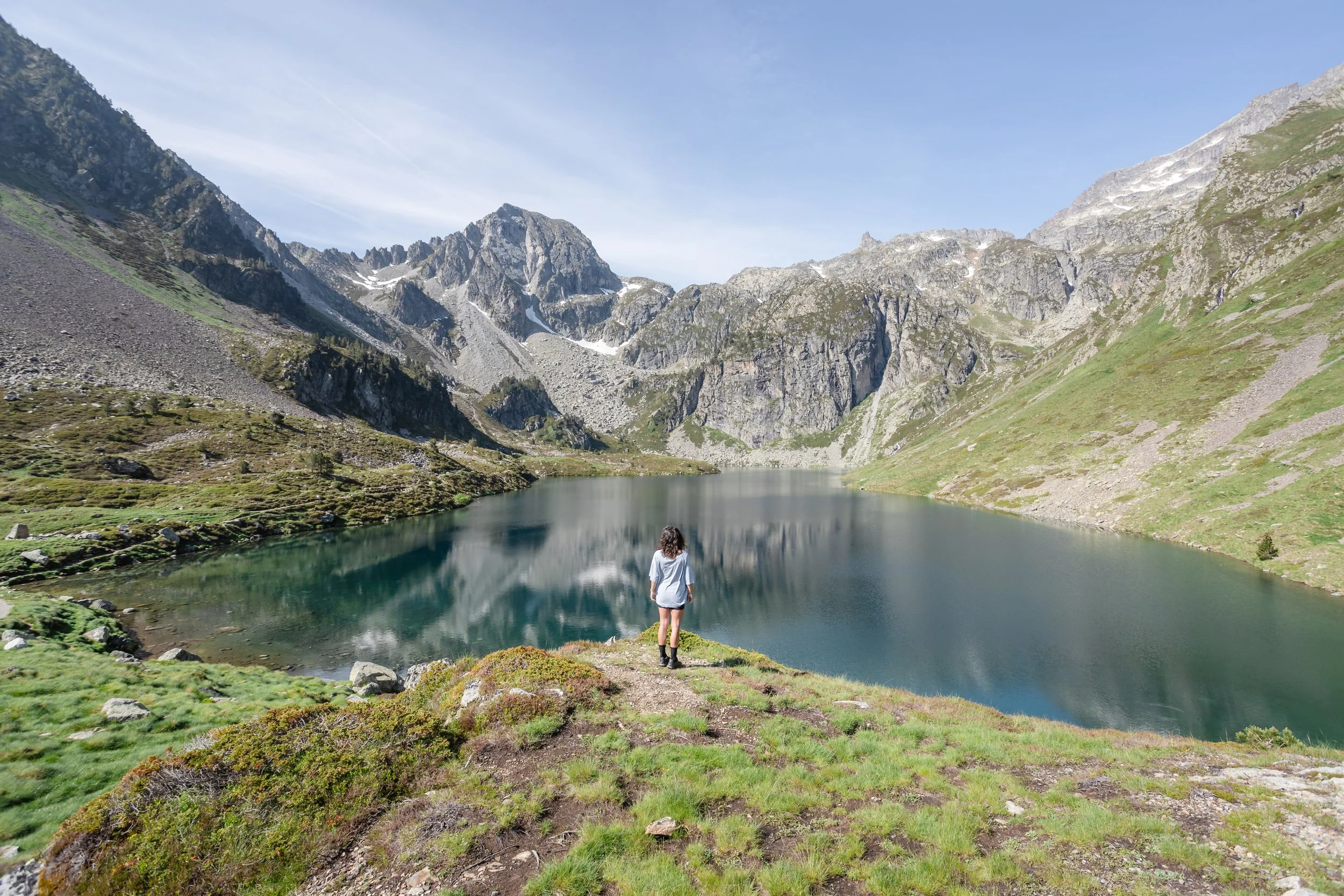 Une femme debout au bord d’un lac de montagne entouré de sommets rocheux, avec un ciel clair et quelques nuages.