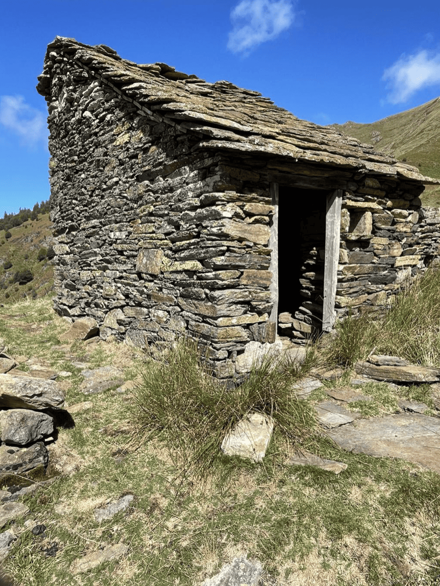 Une petite cabane en pierre dans un paysage de montagne avec un ciel bleu et quelques nuages.