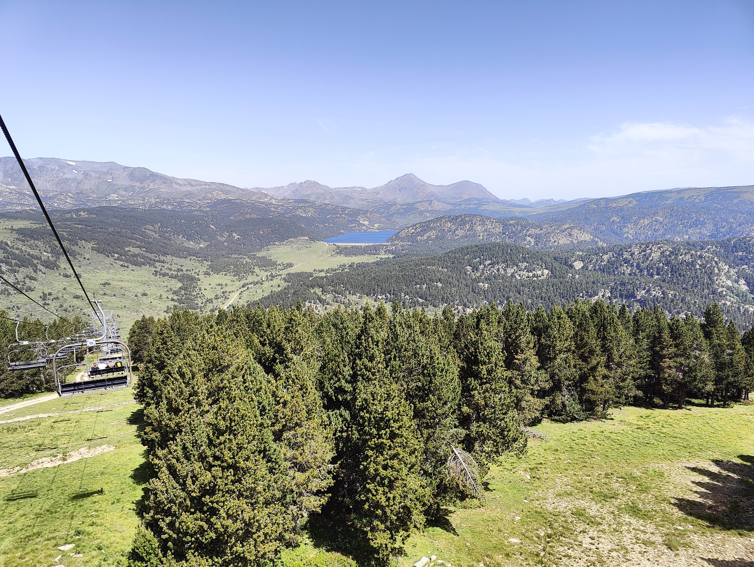 Vue panoramique d'une étendue de nature avec des montagnes, un lac, des forêts et un téléphérique en déplacement.