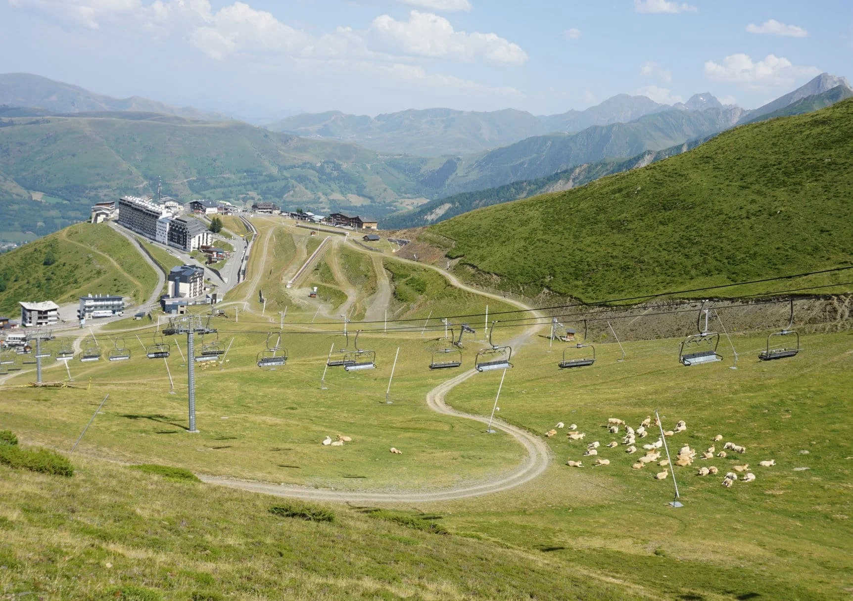 Une station de ski en montagne avec des télécabines, des bâtiments, un pâturage avec des moutons, et un paysage de montagnes vertes derrière.