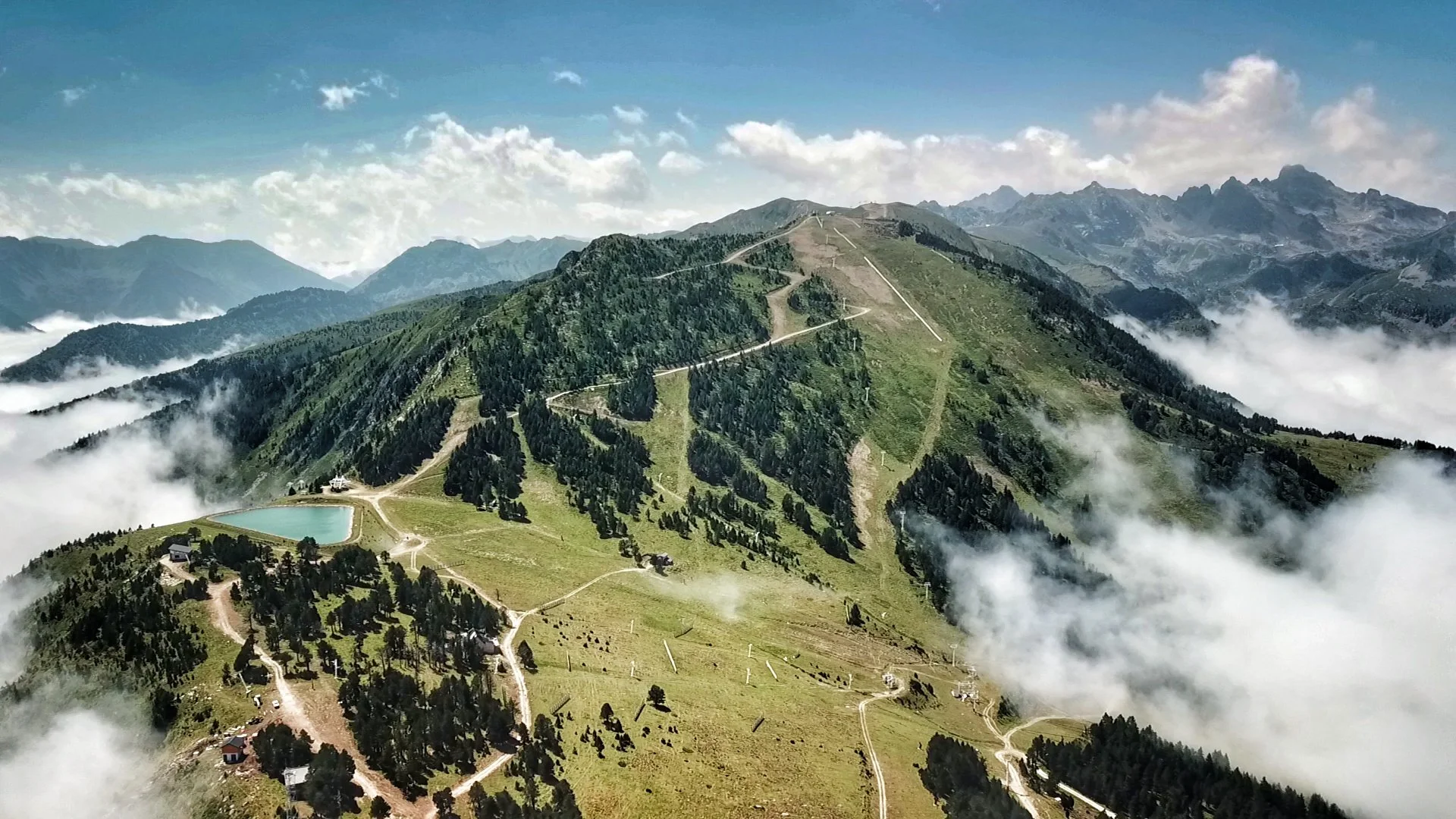 Montagne verdoyante avec des sentiers, un lac, des arbres dispersés, nuages en altitude et montagnes rocheuses en arrière-plan.