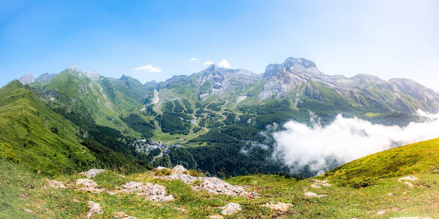 Vue panoramique sur une vallée verdoyante entourée de montagnes avec des sommets rocheux, un petit village au fond et des nuages au sol.