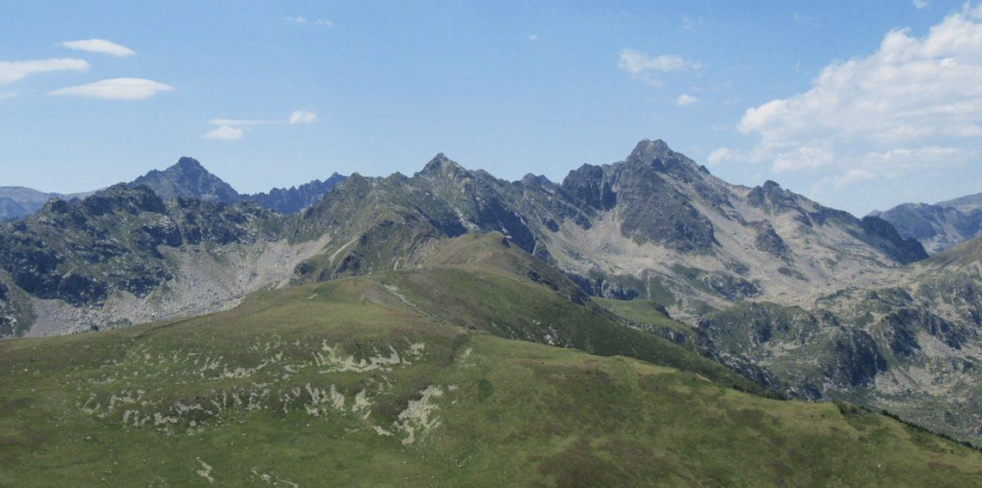 Paysage de montagnes rocheuses avec en premier plan une colline verte et un ciel bleu avec quelques nuages.