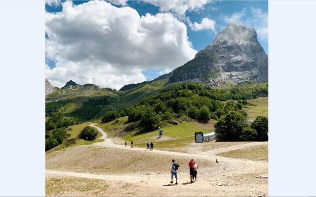 Paysage montagneux avec sentiers, quelques personnes marchant ou prenant des photos, vegetation verte, ciel avec des nuages.