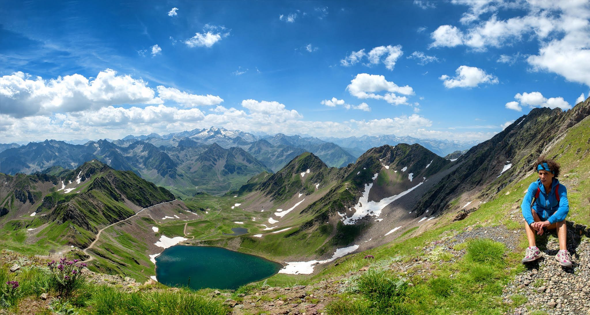 Une femme en vêtements de randonnée assise sur un rocher dans un paysage de montagnes verdoyantes avec un lac alpin, sous un ciel bleu ponctué de nuages.