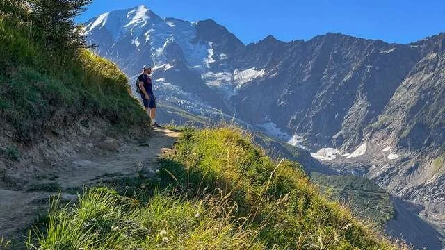 Homme marchant sur un sentier de montagne avec un paysage de sommets enneigés et de vallées verdoyantes en arrière-plan.