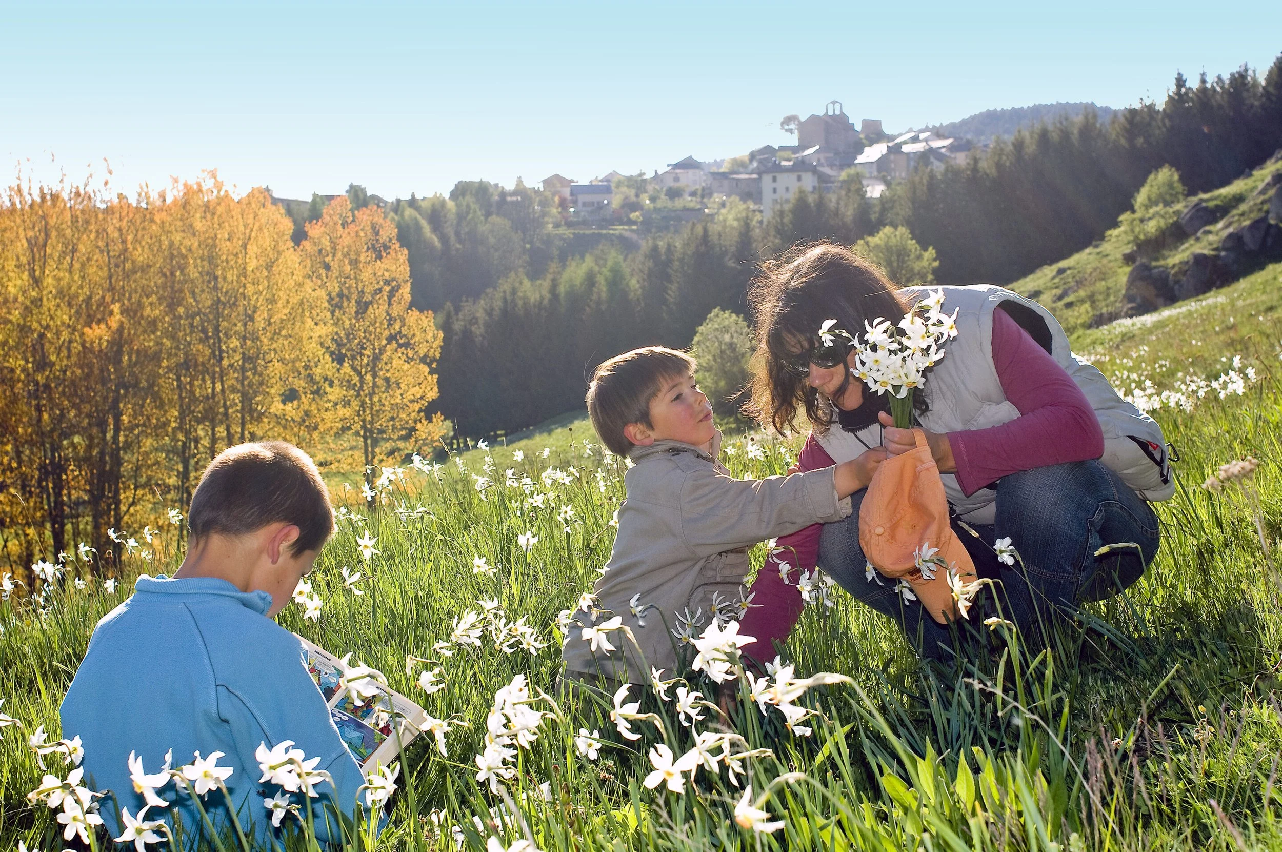 Une femme et deux enfants dans un champ de fleurs sauvages en montagne, avec des arbres aux couleurs d'automne et un village en arrière-plan.