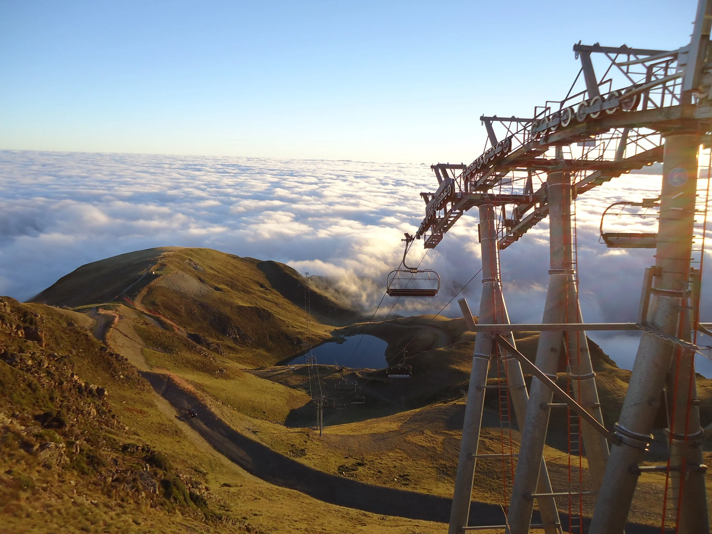Téléphériques suspendus au-dessus de collines verdoyantes avec une mer de nuages en arrière-plan, au soleil couchant.