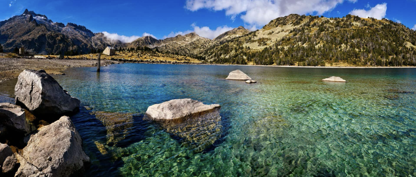 Lac clair avec des rochers, entouré de montagnes rocheuses et boisées, sous un ciel bleu avec quelques nuages.