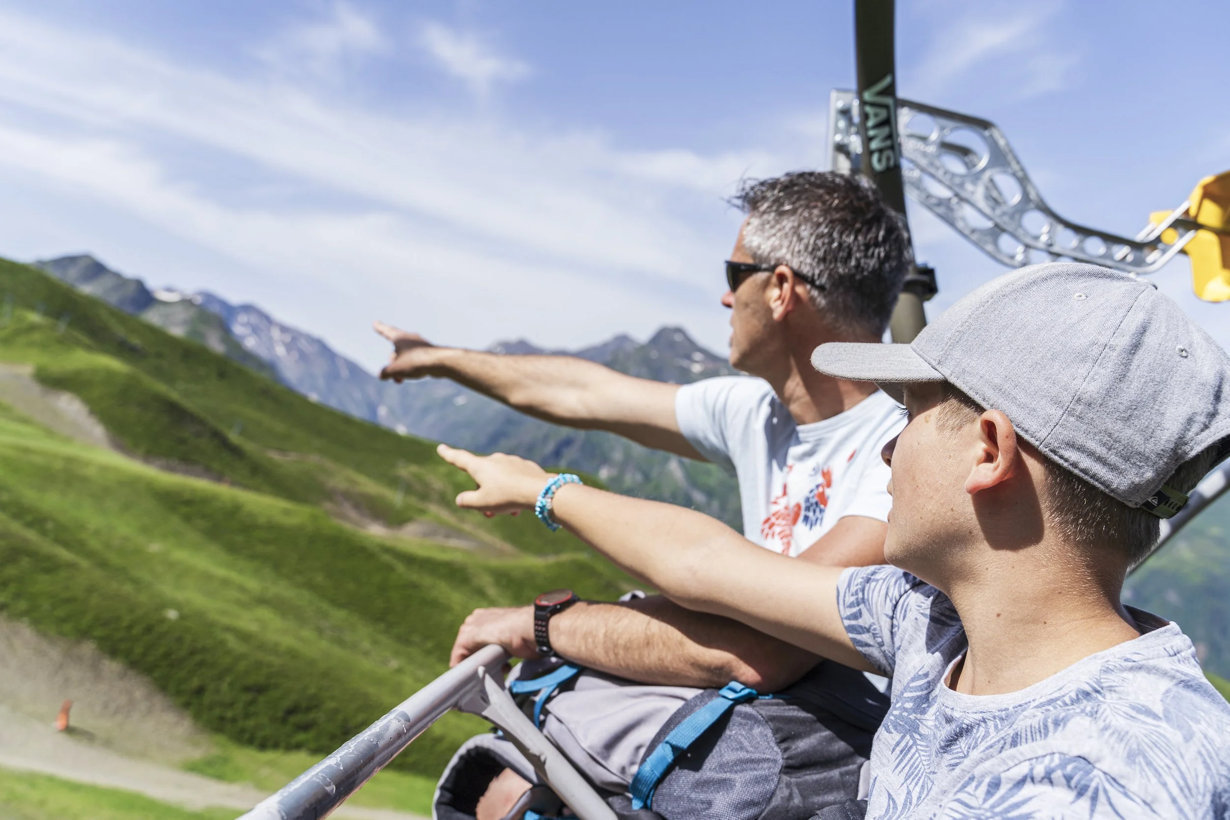 Un homme adulte avec des lunettes et une chemise à motifs, assis avec deux enfants, pointant vers l'horizon dans un paysage de montagnes verdoyantes sous un ciel bleu clair.