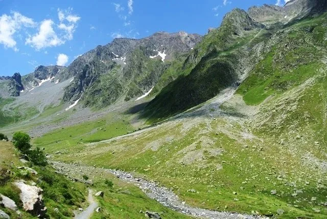 Paysage de montagne avec des sommets rocheux, des pentes vertes et un ruisseau au premier plan, sous un ciel bleu avec quelques nuages.