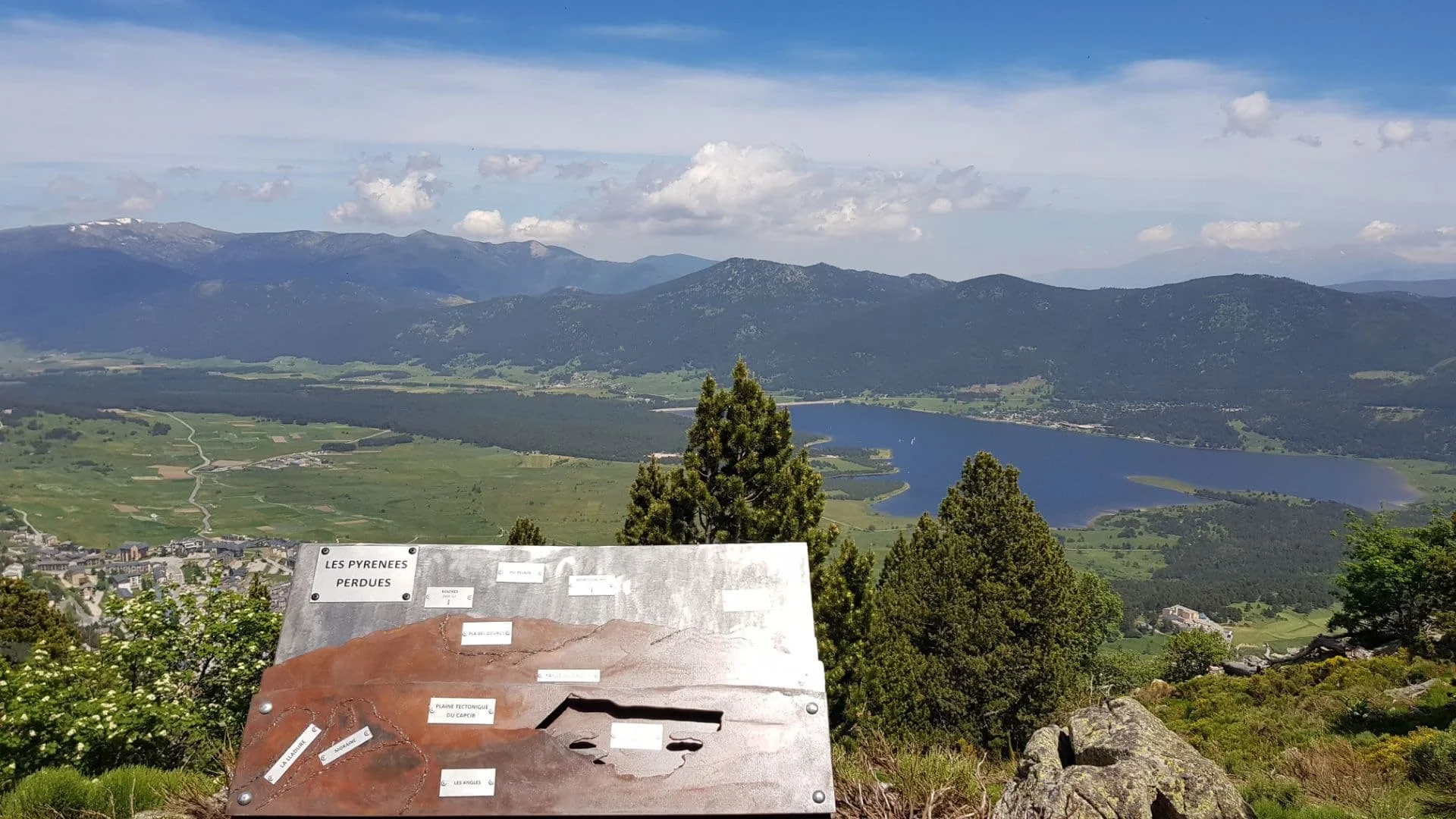 Vue panoramique sur les montagnes des Pyrénées, un lac, des forêts, un village, et un panneau explicatif en premier plan.