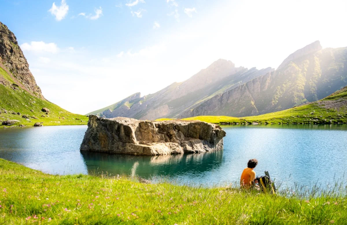Personne assise sur l'herbe près d'un lac entouré de montagnes, avec un gros rocher au centre du lac, en plein jour.