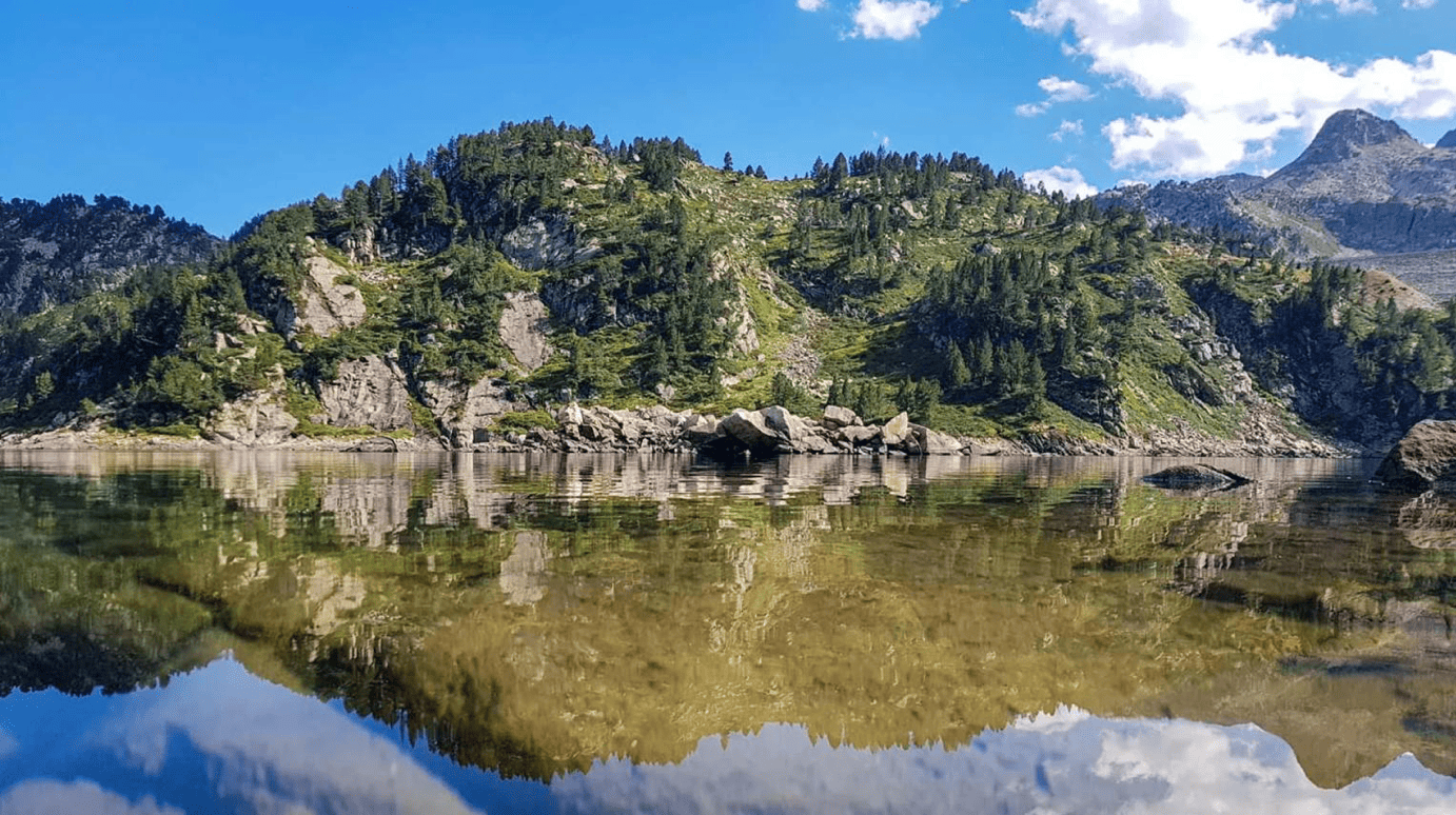 Paysage de montagne avec des forêts verdoyantes et un lac reflétant la nature, sous un ciel bleu avec quelques nuages.