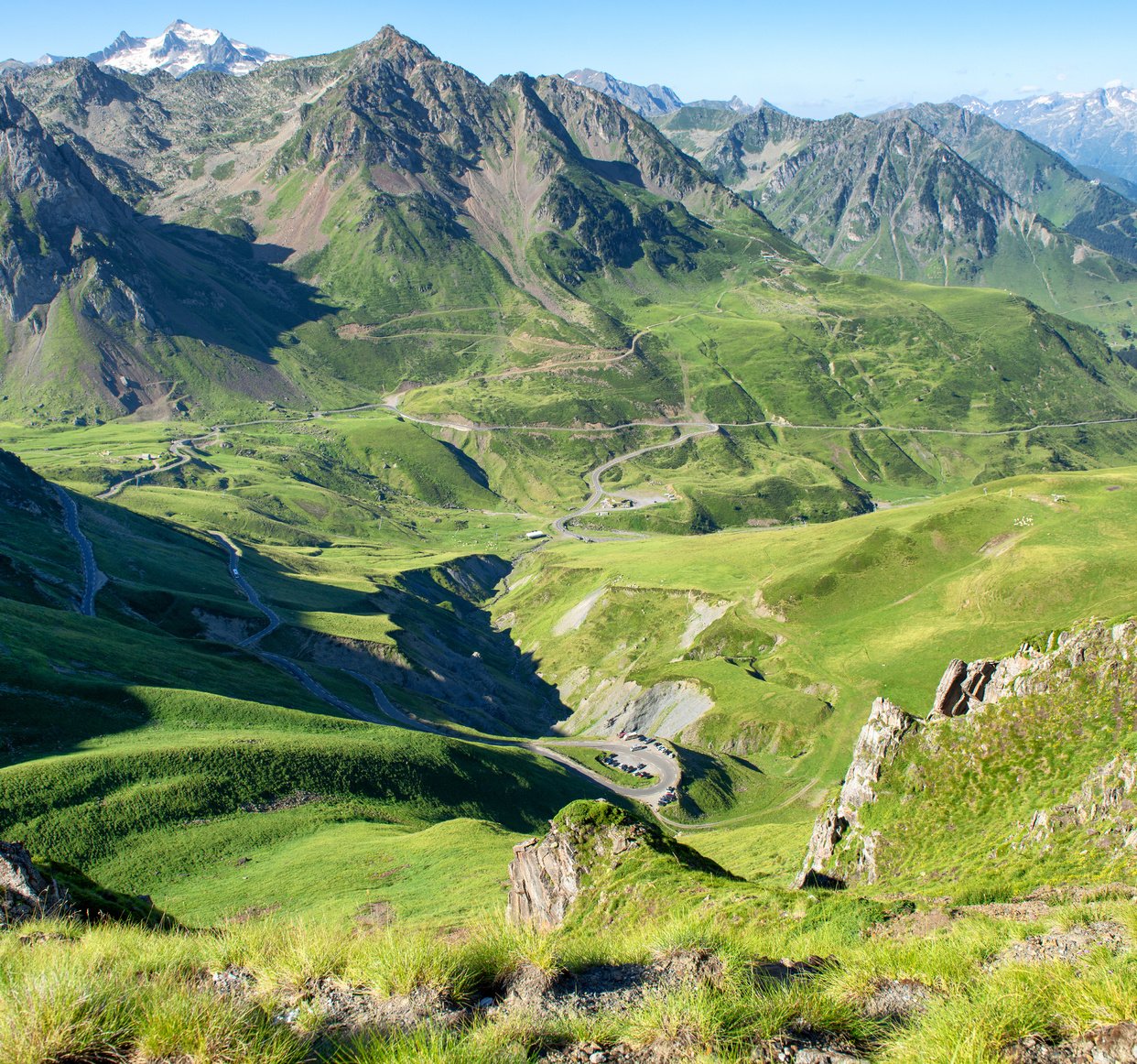 Paysage de montagnes verdoyantes avec routes serpentines, vallées et sommets enneigés en arrière-plan