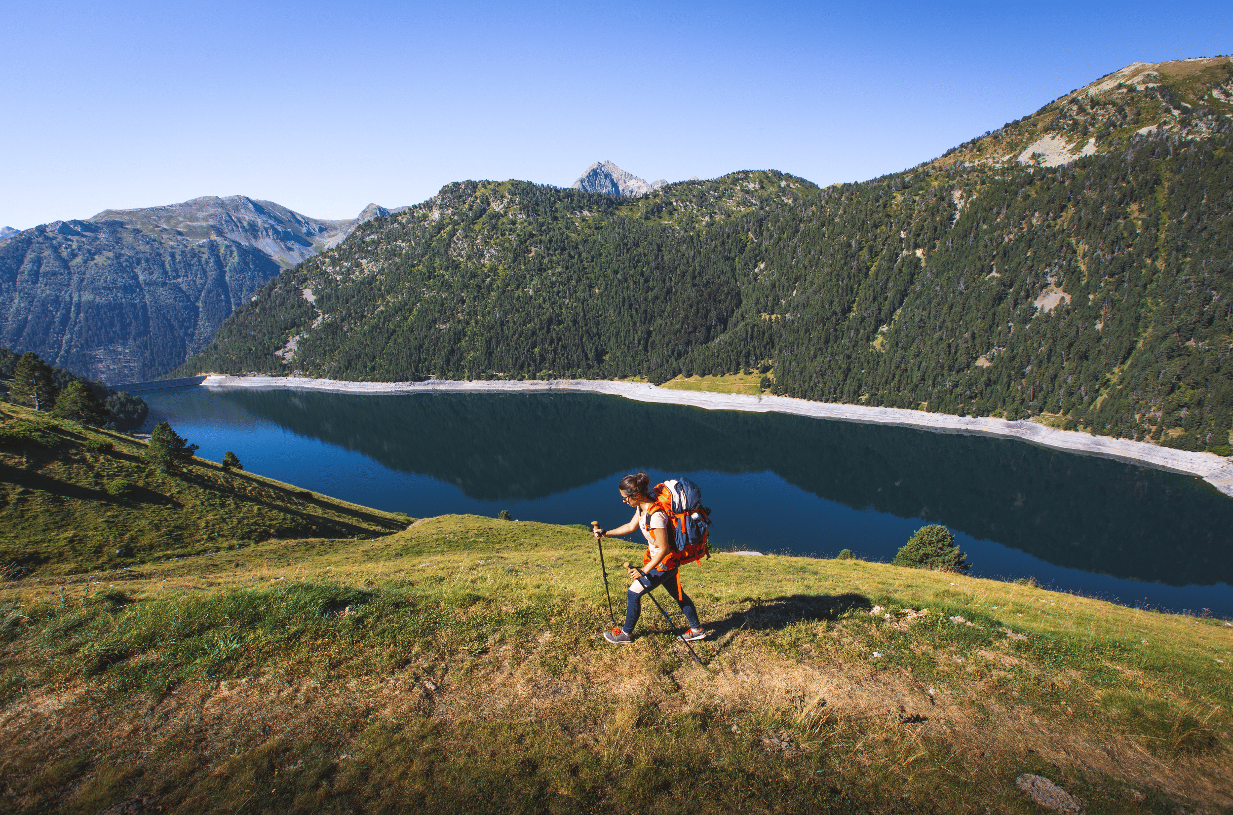 Une randonneuse équipée de bâtons, portant un sac à dos, marche sur une colline herbeuse près d'un lac en montagne avec des paysages de montagnes et une forêt dense en arrière-plan.