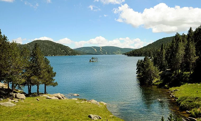 Lac entouré d'arbres avec des montagnes en arrière-plan, ciel bleu avec quelques nuages.