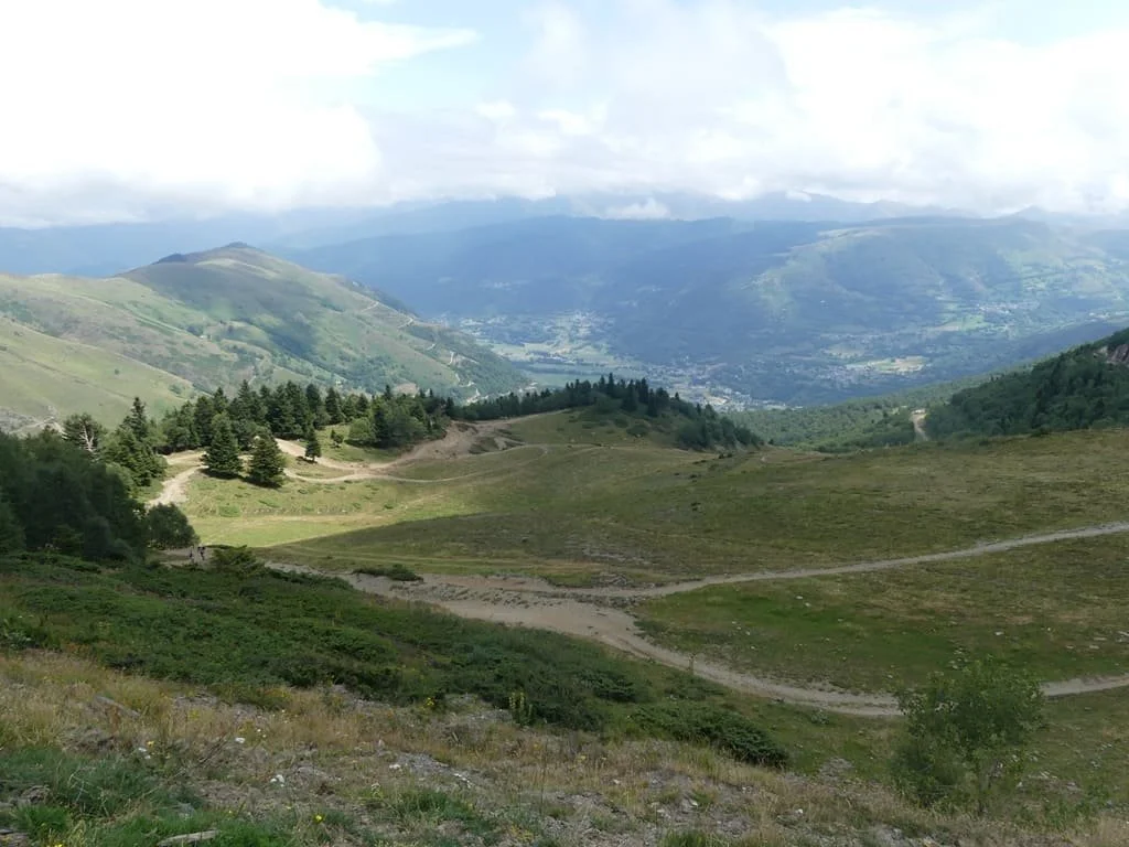 Paysage de montagnes verdoyantes avec des sentiers sinueux, des arbres dispersés et un ciel nuageux.