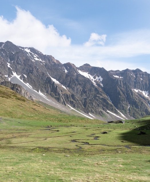 Vallée verdoyante entourée de montagnes rocheuses avec quelques traces de neige et un ciel partiellement nuageux.