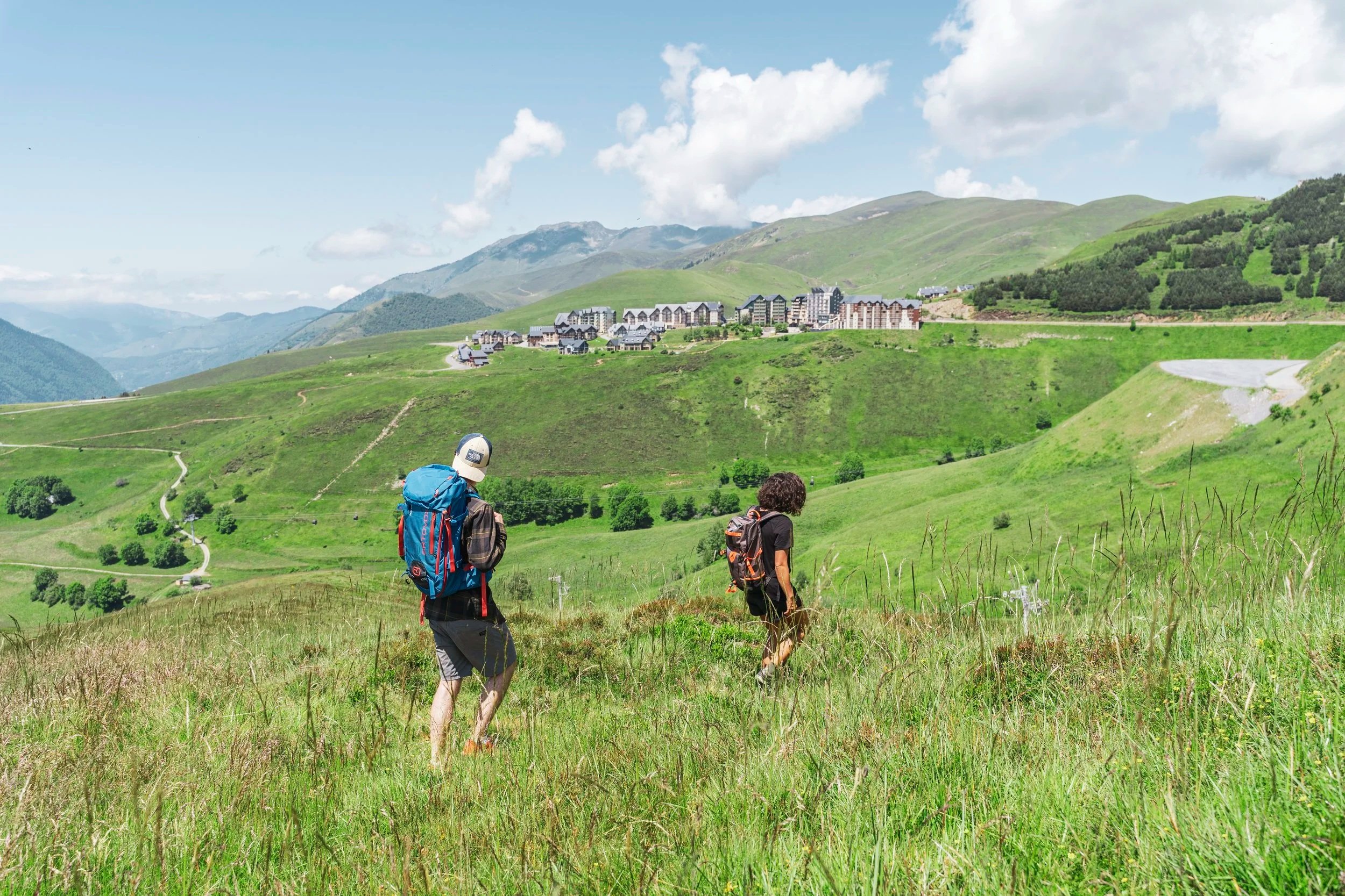 Deux randonneurs marchent dans une prairie verte avec des montagnes en arrière-plan et un village en contrebas.